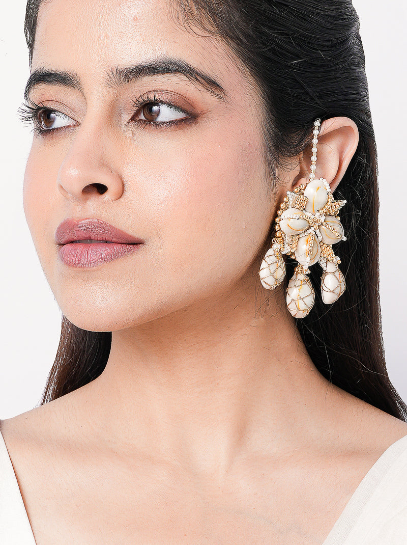 Charvi Floral Shell Earrings featuring cowrie shell floral motifs, faux pearl detailing, ear chain support and a gold tone finish for bridal and festive wear.