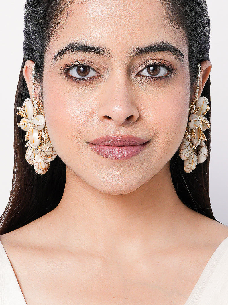 Charvi Floral Shell Earrings featuring cowrie shell floral motifs, faux pearl detailing, ear chain support and a gold tone finish for bridal and festive wear.